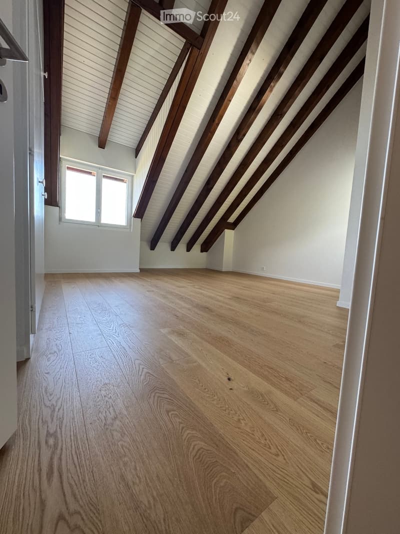 Empty attic space with wooden beams, light wooden flooring, and a small window letting in natural light.