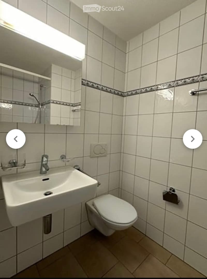 A small bathroom with white tiled walls and floor, featuring a sink, toilet, mirror, and showerhead.