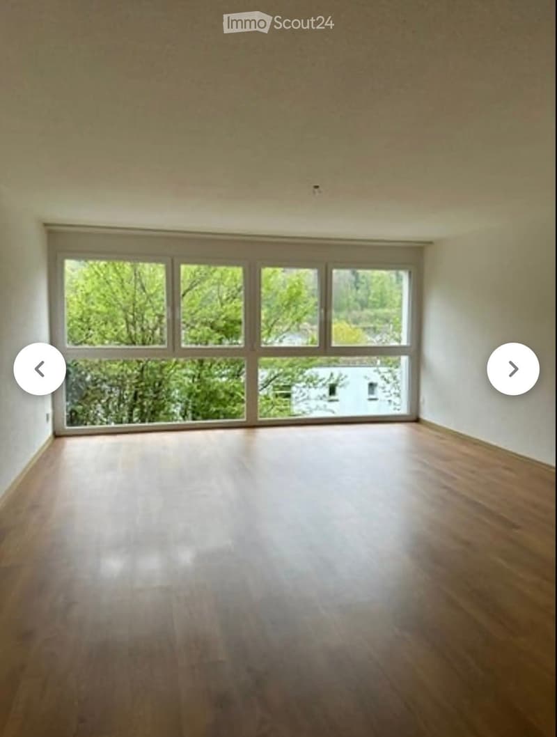 Empty room with wooden flooring and large windows showing greenery outside.