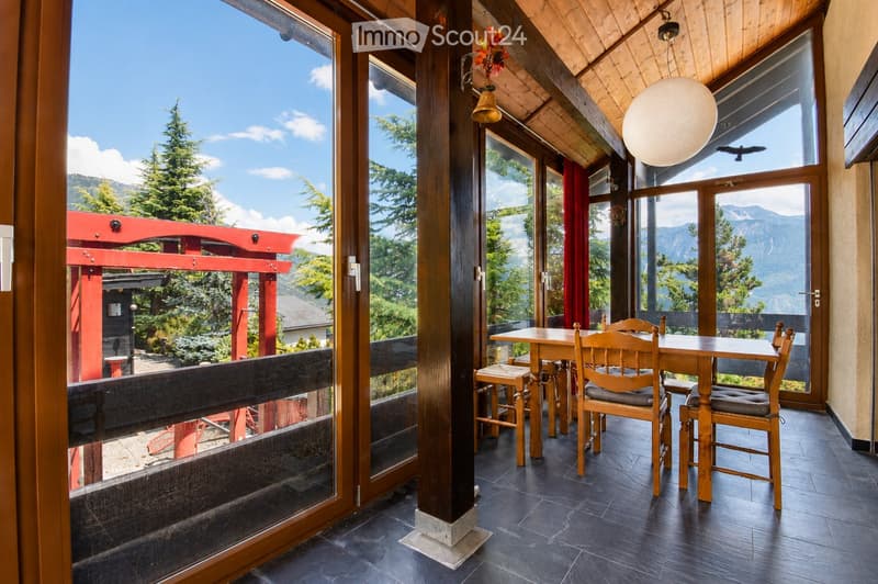 Bright, wooden-framed balcony with large windows offering mountain views, furnished with a wooden table and chairs.
