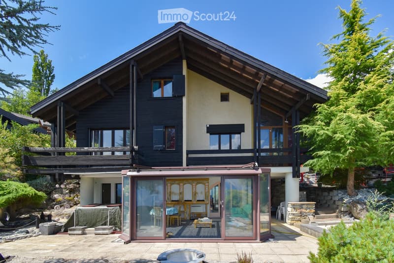 A two-story house with a dark wooden exterior and light-colored walls, featuring large windows and a balcony. The house is surrounded by greenery and includes an outdoor patio area with a glass-enclosed section containing furniture and a small pool.