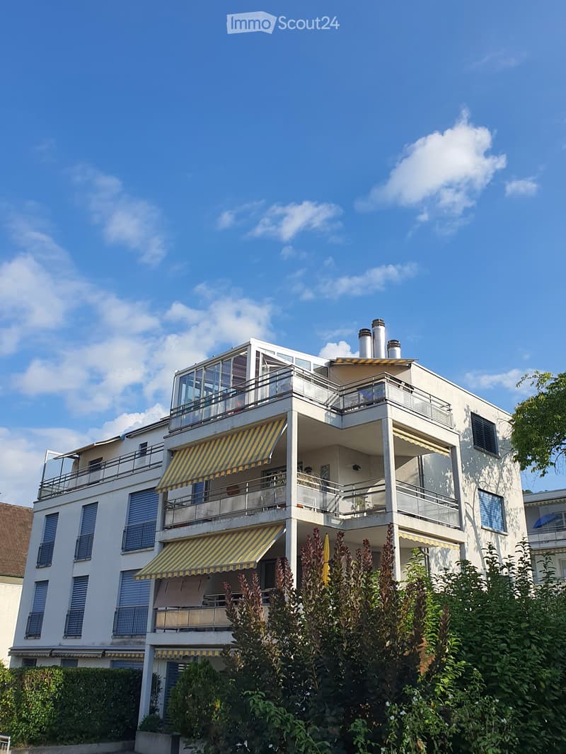 Modern three-story building with white facade, multiple balconies, striped awnings, and visible chimneys on the roof.