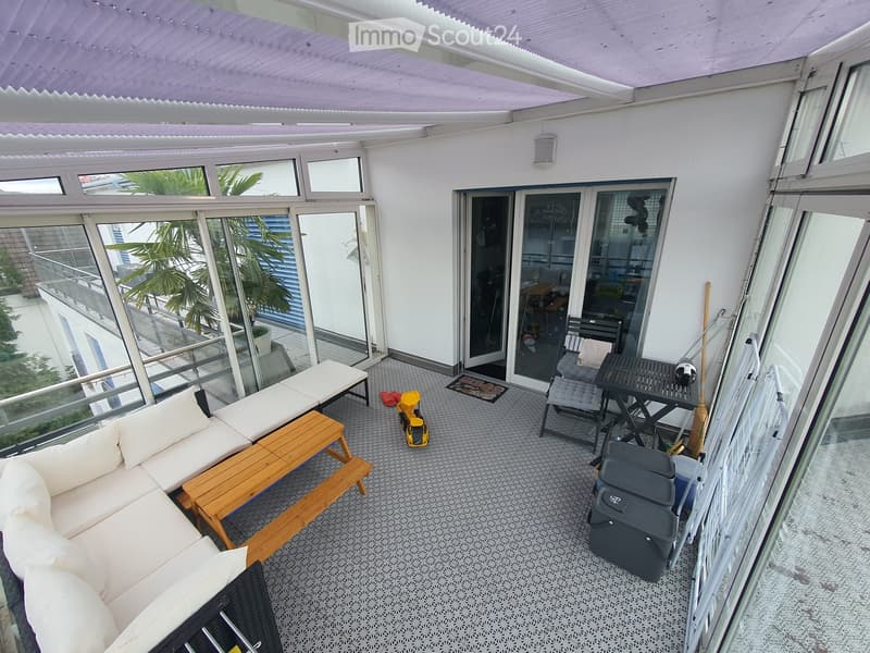 Balcony with a white sofa, wooden table, chairs, and a small stool, with large windows and a view of greenery.