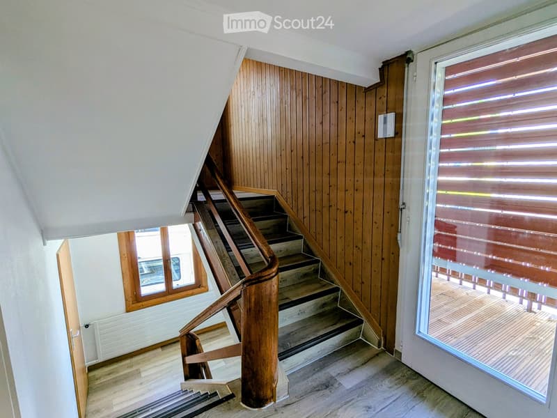 Wooden staircase with a curved handrail leads to an upper level, flanked by wooden panel walls and a large window with blinds. The floor is light-colored wood, and the ceiling is white with a sloped design.