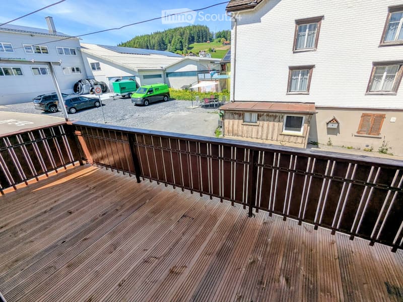 Wooden balcony with metal railing, overlooking a parking area with several vehicles and buildings in the background.