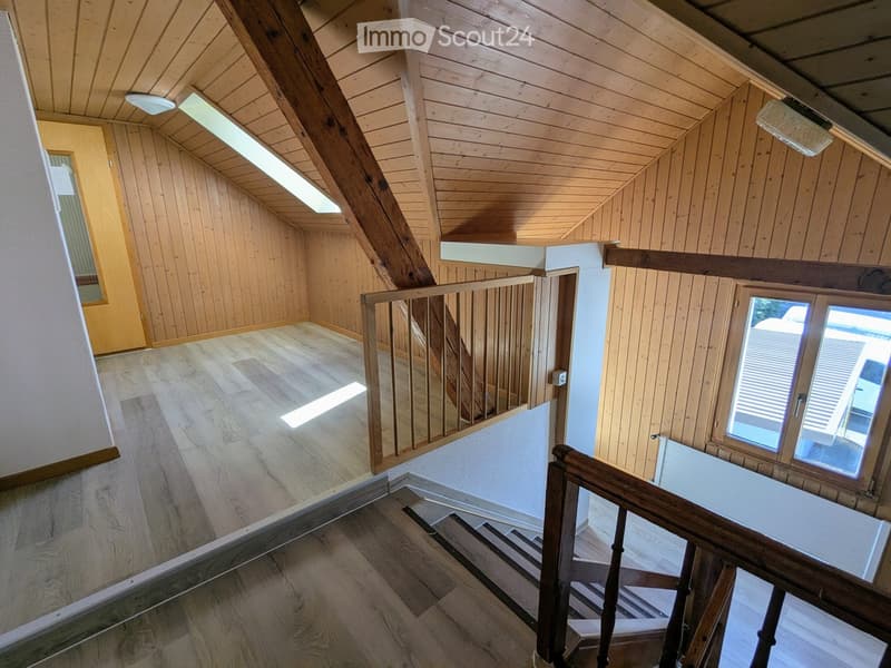 Wooden staircase with wooden railings, wooden walls, and ceiling beams, leading to an open loft area with skylights and large windows.