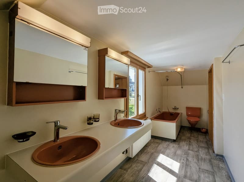 Modern bathroom with two sinks, a bathtub, a toilet, and a large window providing natural light. Features include wooden shelves, a built-in mirror, and a sleek faucet design.
