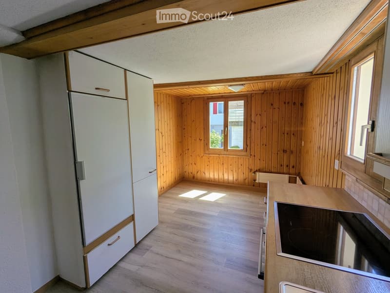 Modern kitchen with light wooden cabinets, a refrigerator, a stove, and a sink. The space features wooden walls and flooring with large windows allowing natural light.