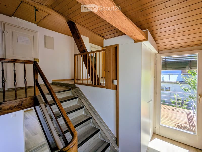 Wooden staircase with wooden handrail and balustrade, leading to an open wooden ceiling area. White walls, wooden floor, and a door leading to a balcony with outdoor view.