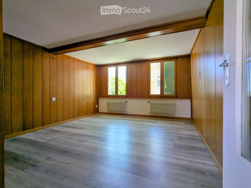 Empty room with wooden walls, light wooden flooring, and large windows with shutters. Radiators are visible under the windows.