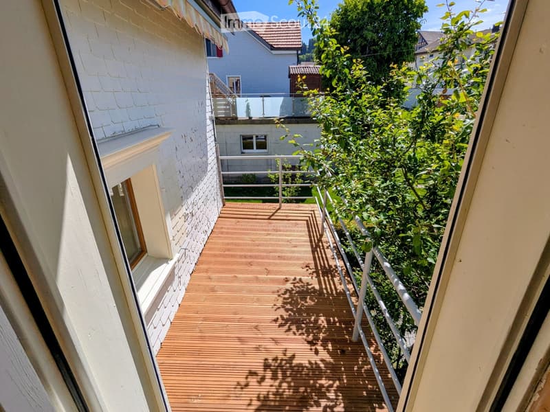 Wooden balcony with white railing, surrounded by greenery and neighboring buildings.
