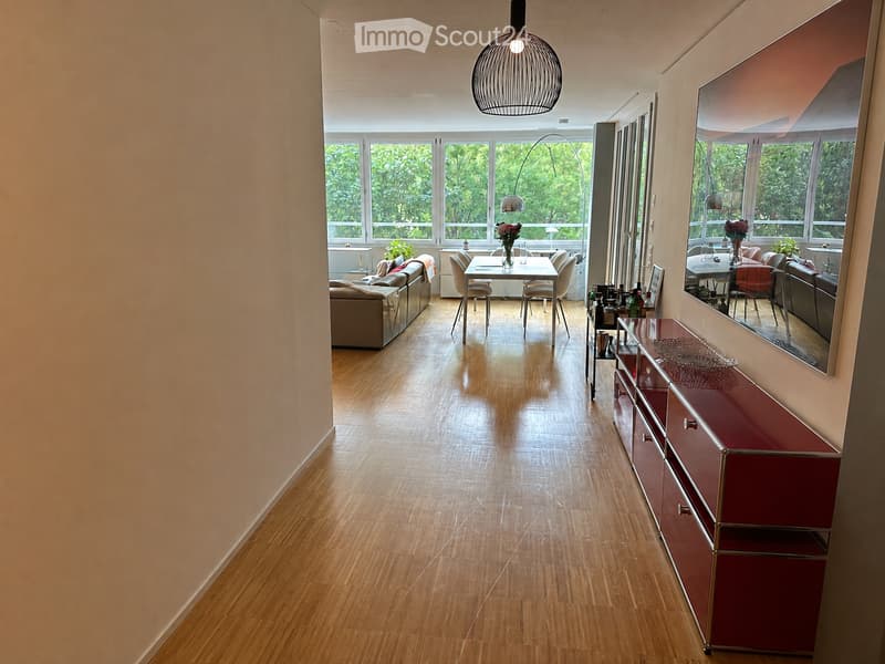 Open-plan living and dining area with wooden flooring, large windows, a sofa, dining table, chairs, a mirror, and a red cabinet.