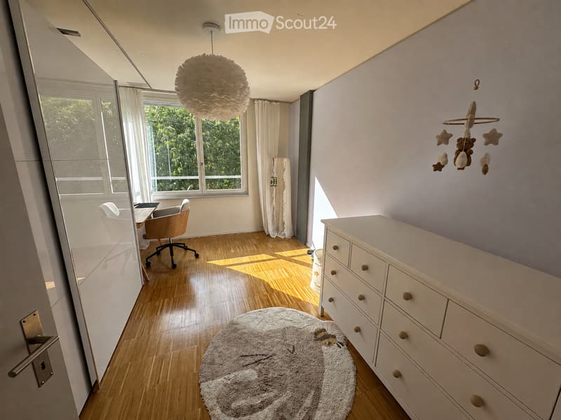 Bright, modern bedroom with wooden flooring, large windows, a white dresser, a round shaggy rug, a hanging baby mobile, and a small workspace area with a chair and table.