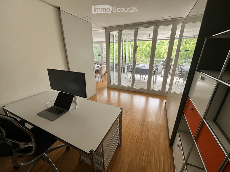 Modern office space with a white desk, laptop, and ergonomic chair. Large glass doors open to a patio area with outdoor furniture.