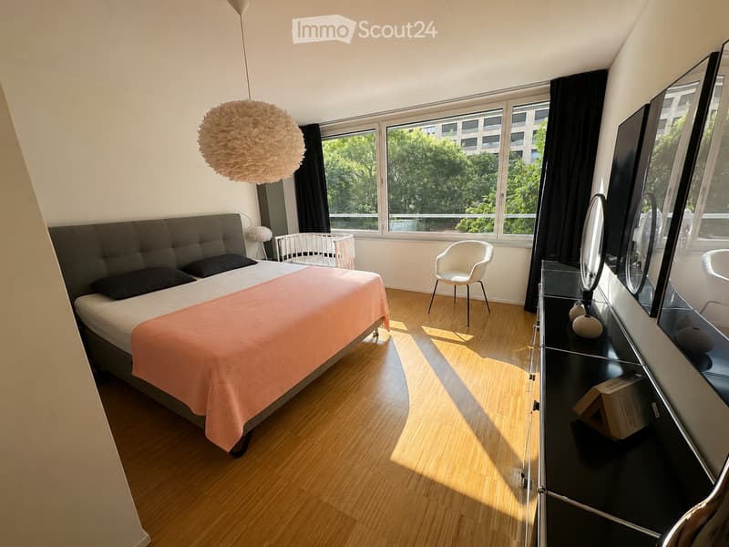 Modern bedroom with wooden flooring, large windows, a bed with a pink blanket, a white chair, a mirror, and a baby crib.