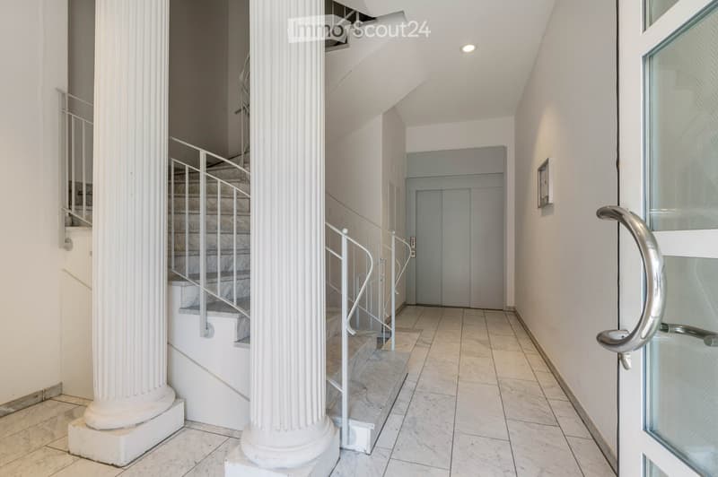 A corridor with marble flooring, white walls, and two large white columns. A staircase with marble steps and metal handrails leads to the upper floor. A door with a glass panel and a silver handle is on the right side.