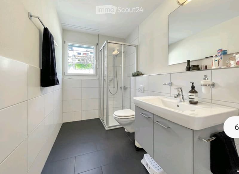 Modern bathroom with white tiles, a glass-enclosed shower, a toilet, a sink with a modern faucet, and a mirror above the sink. A towel hangs on a rack, and a small window provides natural light.