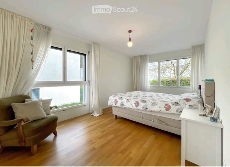 Bright bedroom with wooden flooring, large windows with curtains, a double bed with a patterned duvet, a green armchair, and a nightstand with a lamp.