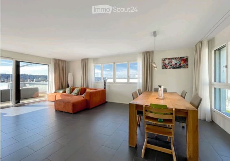 Open-plan living and dining area with large windows offering a view, orange sectional sofa, ottoman, wooden dining table with chairs, and a floor lamp.