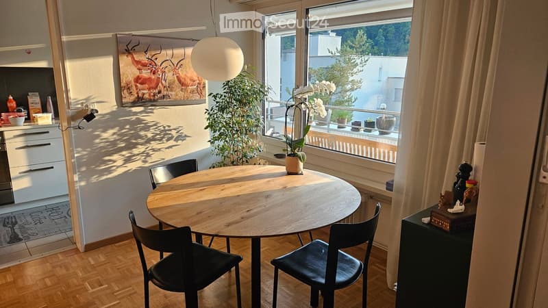 Bright, modern dining area with a round wooden table and black chairs, adjacent to a compact kitchen with white cabinets and a sink. A large window offers a view of the balcony and greenery outside, complemented by a potted plant and a decorative ball lam