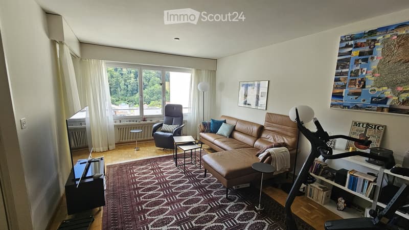Modern living room with large windows, leather sofa, ottoman, armchair, coffee table, side tables, floor lamp, TV, bookshelf, and exercise bike.