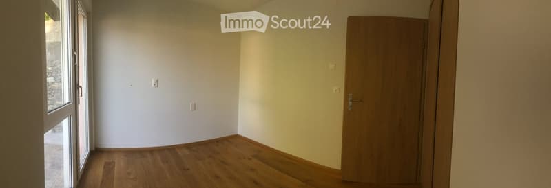 Empty room with wooden flooring, white walls, and a large window with a view of a stone wall outside. A closed wooden door is visible on the right side.