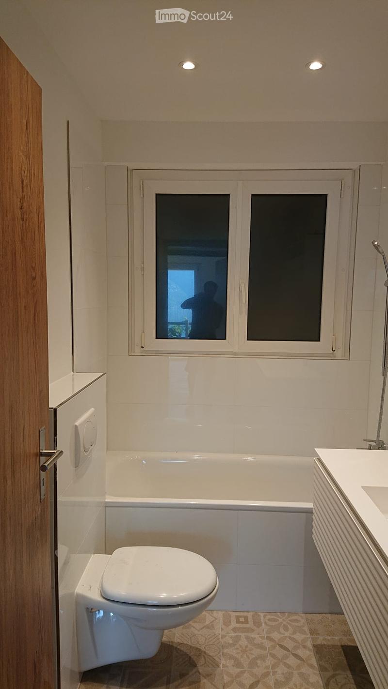 Modern bathroom with a white bathtub, toilet, sink, and a window with frosted glass. The walls are tiled, and the floor has a patterned tile design.