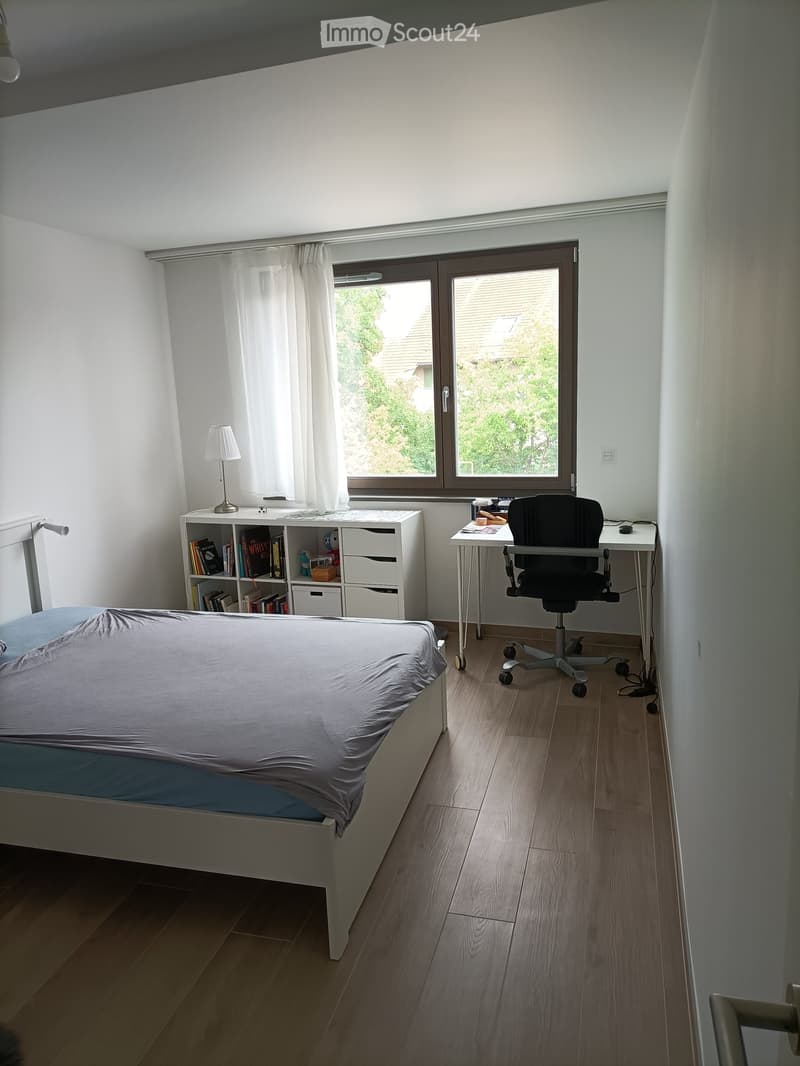 A well-lit bedroom with a bed, desk, chair, bookshelf, and window with curtains.