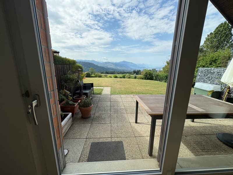 Balcony with tiled floor, wooden table and chairs, potted plants, and a view of a green lawn and distant mountains.