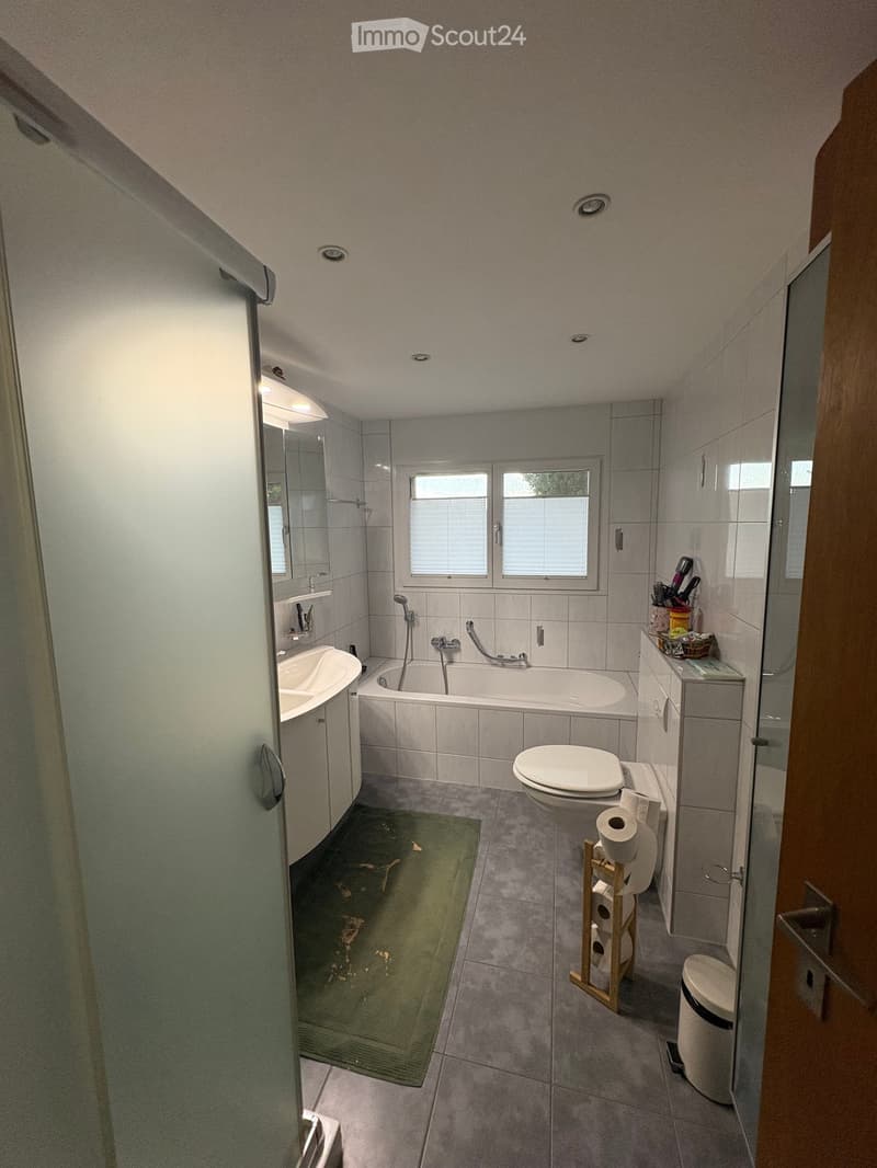 Modern bathroom with a bathtub, toilet, sink, mirror, and a small cabinet. The room features grey tiles, a green bath mat, and a window with blinds.