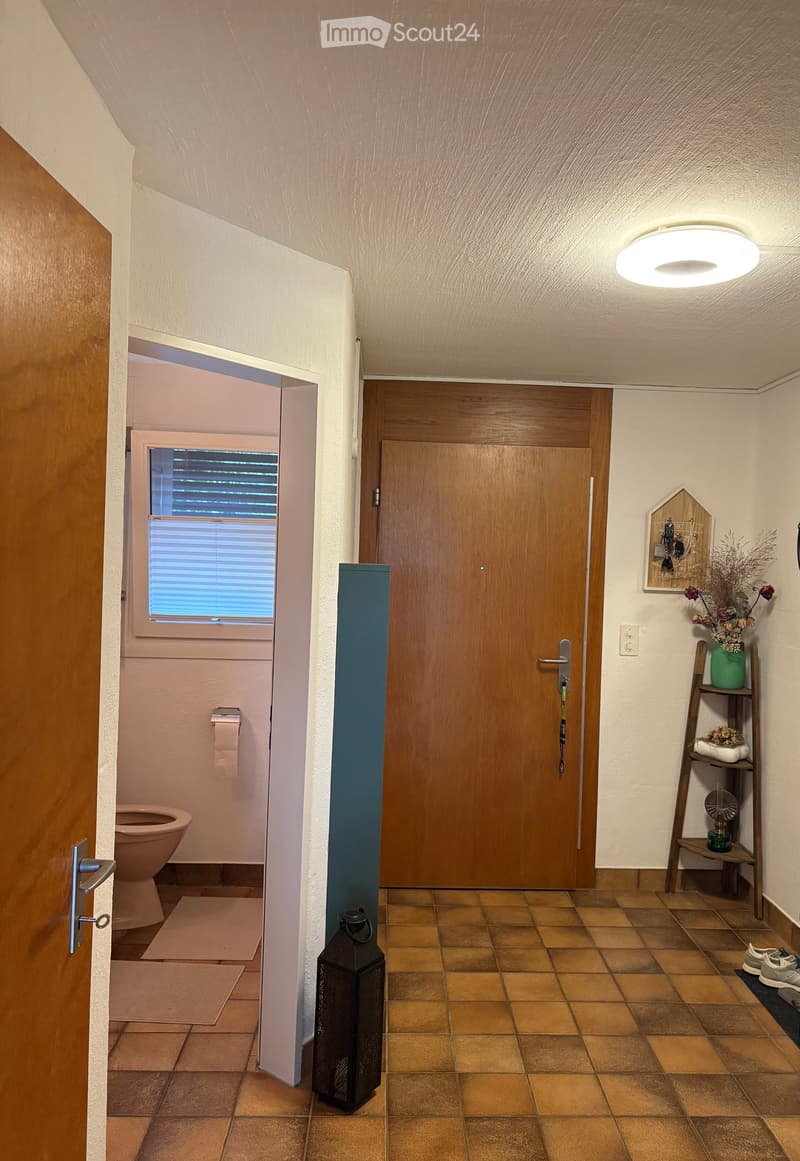 Hallway with tiled floor, wooden doors, a small shelf with decorative items, and a lantern. A toilet is visible through an open door on the left.