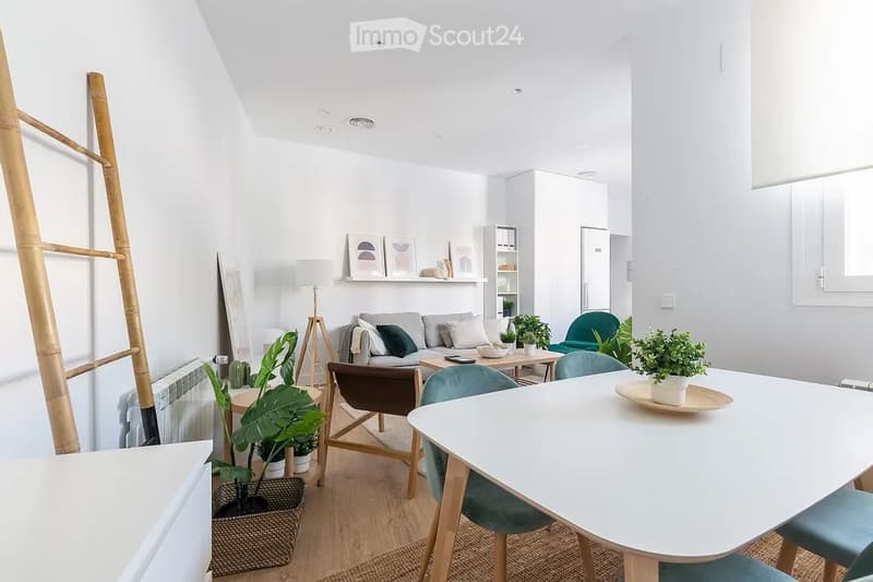 Bright, modern living room with white walls, wooden flooring, and large windows. Features a grey sofa, coffee table, dining table with chairs, potted plants, and a floor lamp.