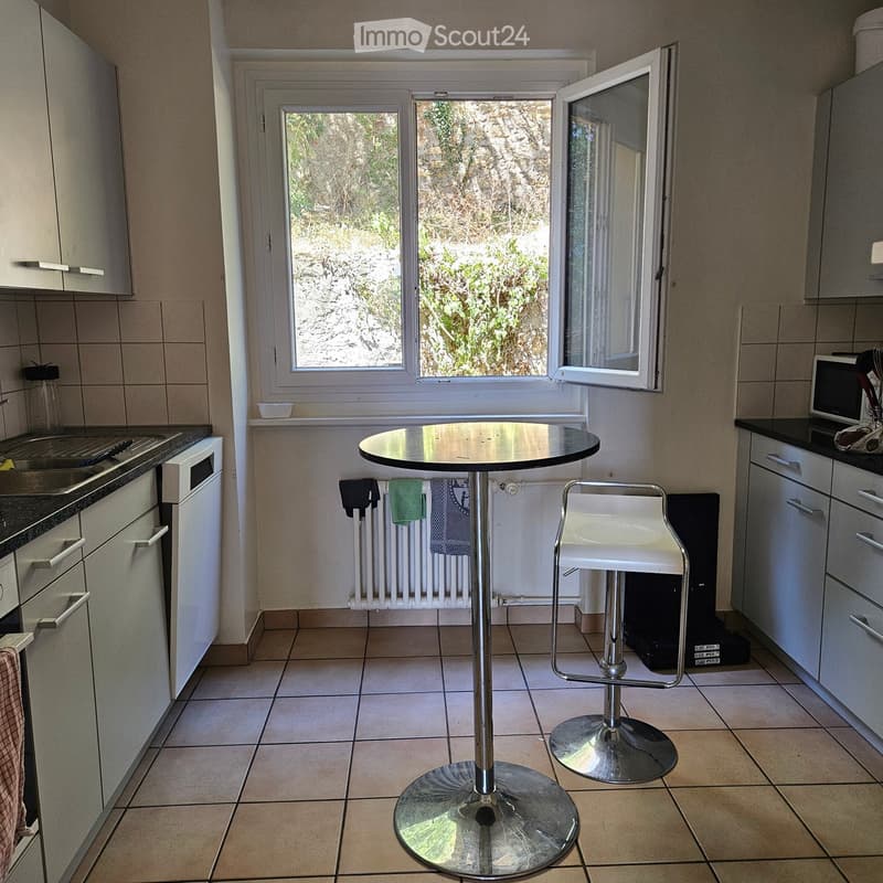 Modern kitchen with tiled floor, white cabinets, stainless steel appliances, a round table with a high stool, and a window overlooking a rocky outdoor area.
