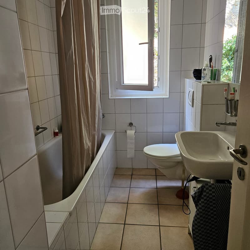 Bathroom with a bathtub, toilet, sink, mirror, and a window with a shower curtain. Toilet paper and a towel are visible.