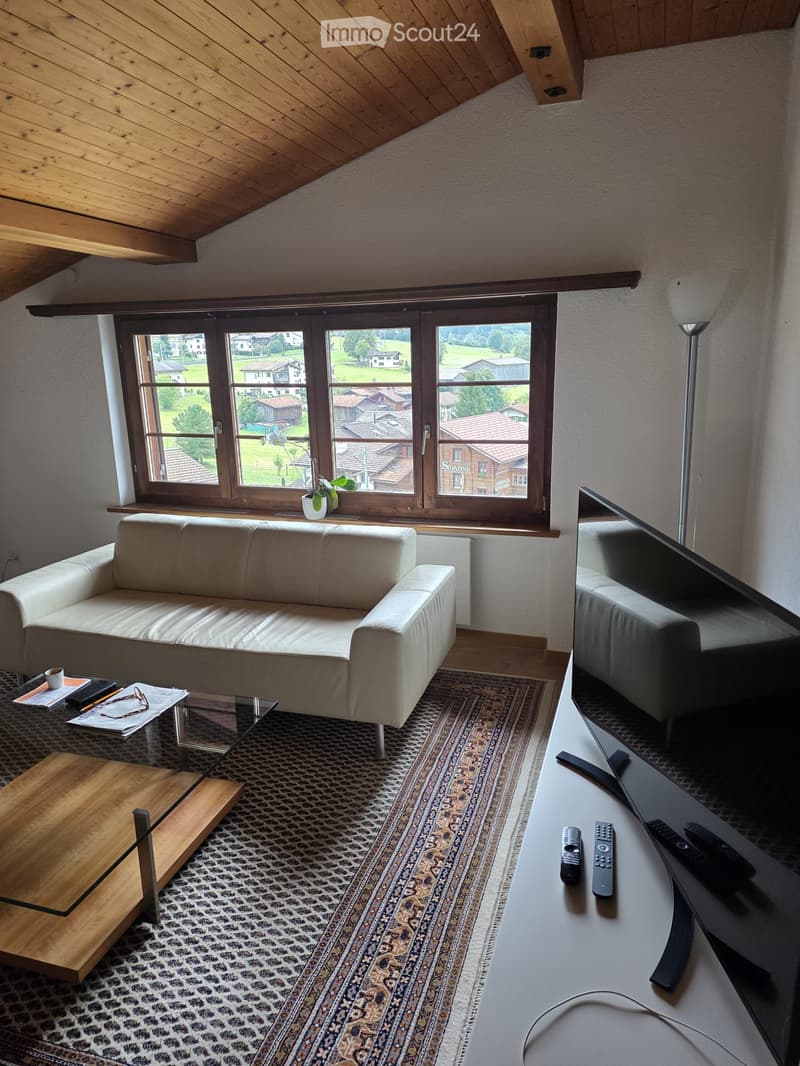 Modern living room with a white leather sofa, glass coffee table, wooden side table, large television, and scenic view through large windows.