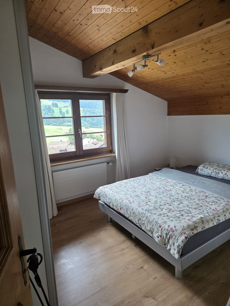 Cozy attic bedroom with wooden slanted ceiling, large window offering scenic view, single bed with floral bedding, radiator, and white curtains.