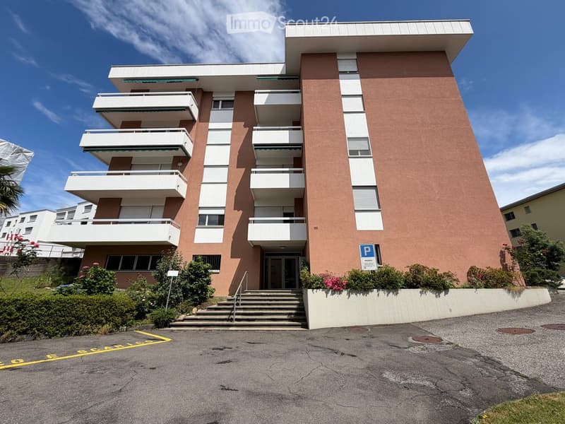 Modern multi-story apartment building with balconies, pink facade, and parking area.