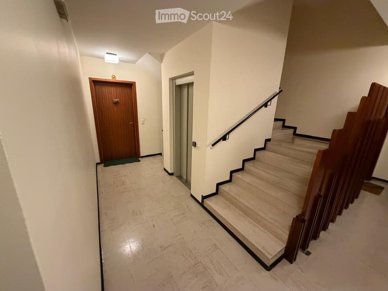 Hallway with tiled floor, wooden door, staircase with wooden railing, and light fixture on the ceiling.