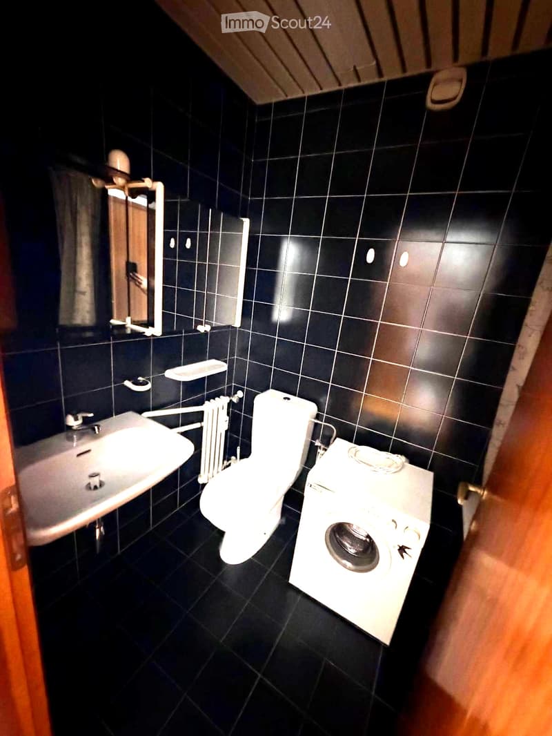 Modern bathroom with black tiled walls and floor, featuring a sink, toilet, radiator, washing machine, and mirror.