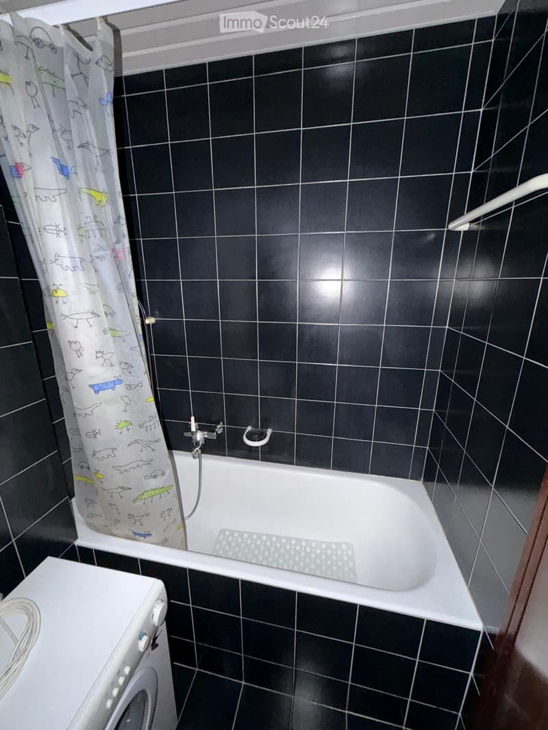 Bathroom with black tiled walls, a white bathtub, a shower curtain with cartoon designs, a washing machine, and a bathtub mat.