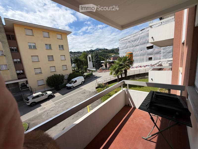 Balcony with tiled floor, metal railing, and a grill on the right side. The view shows a multi-story building, parked cars, and a partially constructed building with scaffolding.