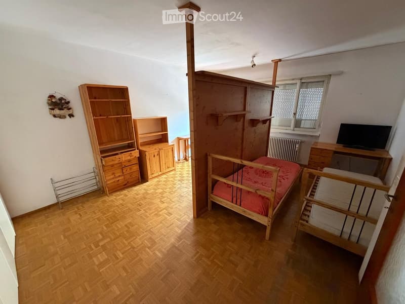 A small bedroom with wooden flooring, a single bed with a red cover, a desk with a screen, a bookshelf, and a radiator near the window.