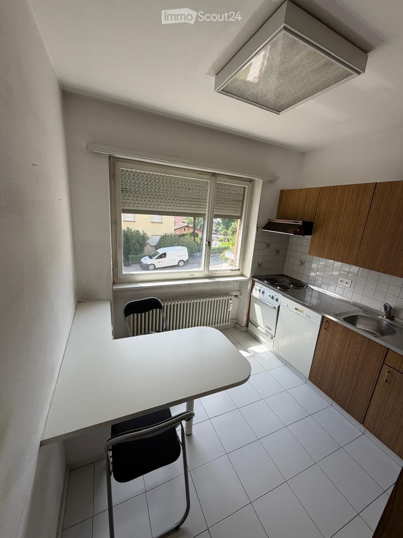 Compact kitchen with white tiled floor, wooden cabinets, stove, oven, dishwasher, sink, and a small table with a chair.