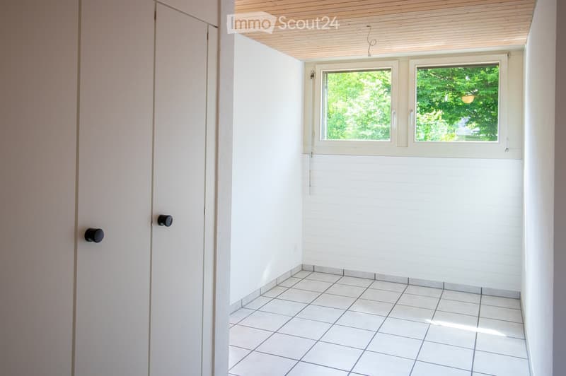 Empty room with white tiled floor, white paneled walls, and two windows showing greenery outside. The room has a simple wooden ceiling and two doors with black handles.