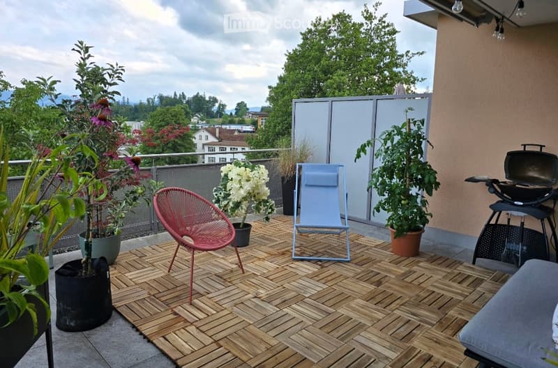 Wooden decked balcony with potted plants, a red chair, a blue lounge chair, and a barbecue grill.