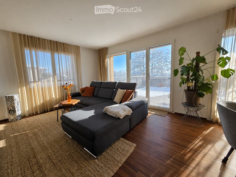 Modern living room with dark grey sectional sofa, wooden coffee table, side tables, potted plant, and large windows with sheer curtains.