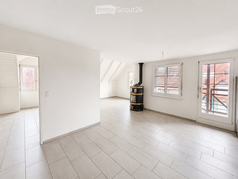 Empty room with white walls, light-colored tiled floor, and a small black wood-burning stove in the center. The room has large windows with blinds and a door leading to a balcony or outdoor area.