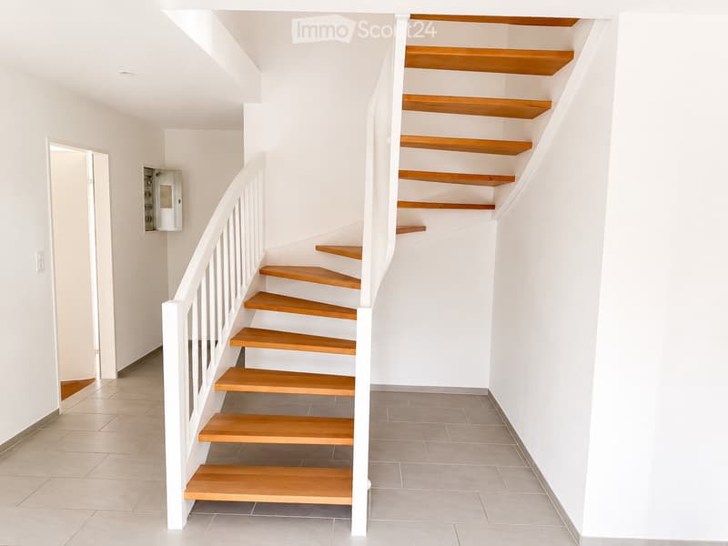 Modern staircase with wooden steps and white railings, leading upwards. The hallway has tiled flooring and white walls, with a small open doorway visible on the left side.