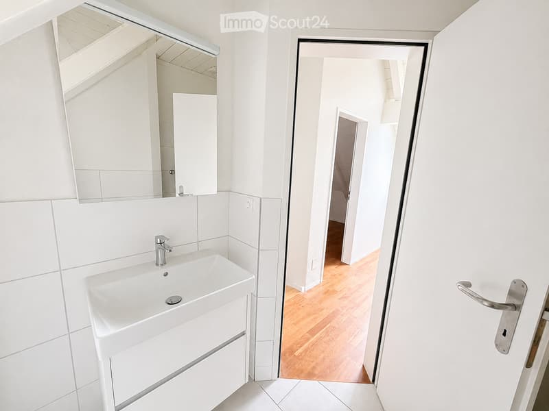 Modern bathroom with white tiles, a white sink, and a large mirror above it. The door leads to another room with wooden flooring.