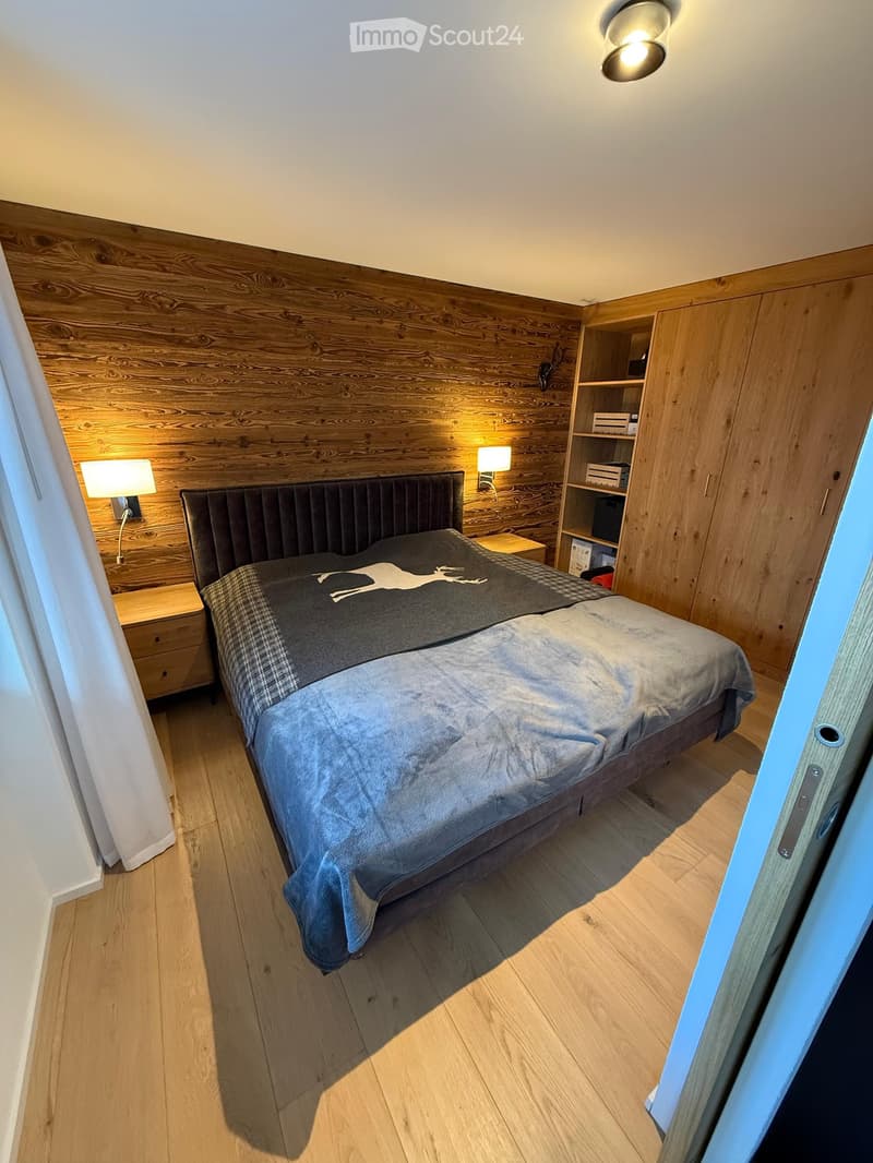 Cozy bedroom with wooden walls, a large bed with a dark headboard, nightstands, wall-mounted lamps, and built-in wooden wardrobe with shelves.